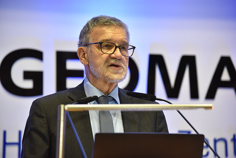 A man with grey hair and a beard speaks at a lectern