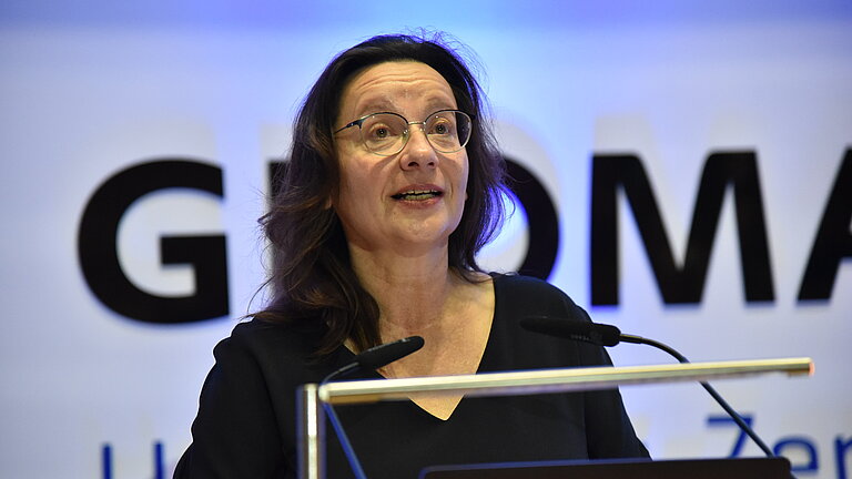 A dark-haired woman in a black dress speaks at a lectern