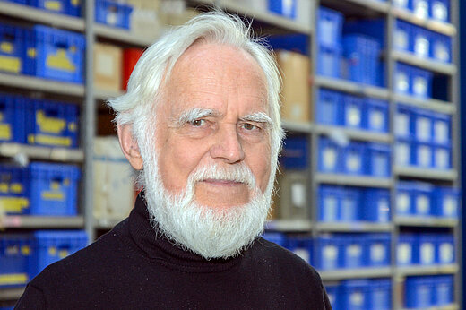 An older man with white hair and a full white beard looks friendly into the camera. High shelves with blue labelled boxes can be seen in the background.