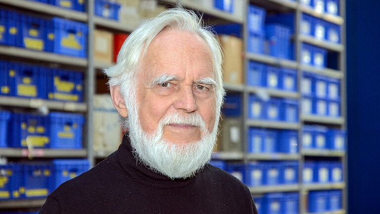 An older man with white hair and a full white beard looks friendly into the camera. High shelves with blue labelled boxes can be seen in the background.