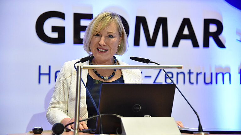 A woman with half-length blonde hair speaks with commitment at a lectern