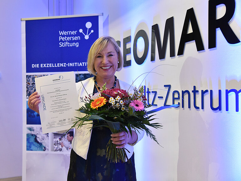 A blonde woman holds a bouquet of flowers and a certificate up to the camera