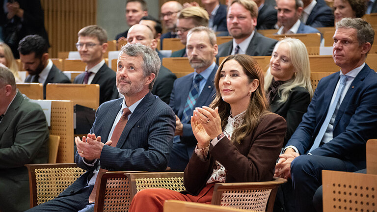 A man and a woman sit on wicker chairs in front of the ascending rows of seats in a lecture theatre that is full to capacity