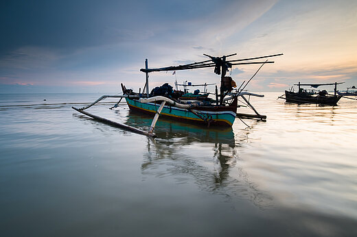 A colourfully painted wooden boat with wooden outriggers is moored on a smooth sea