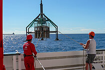  Two workers wearing red hard hats stand on board a research vessel while a piece of research equipment is hoisted on board