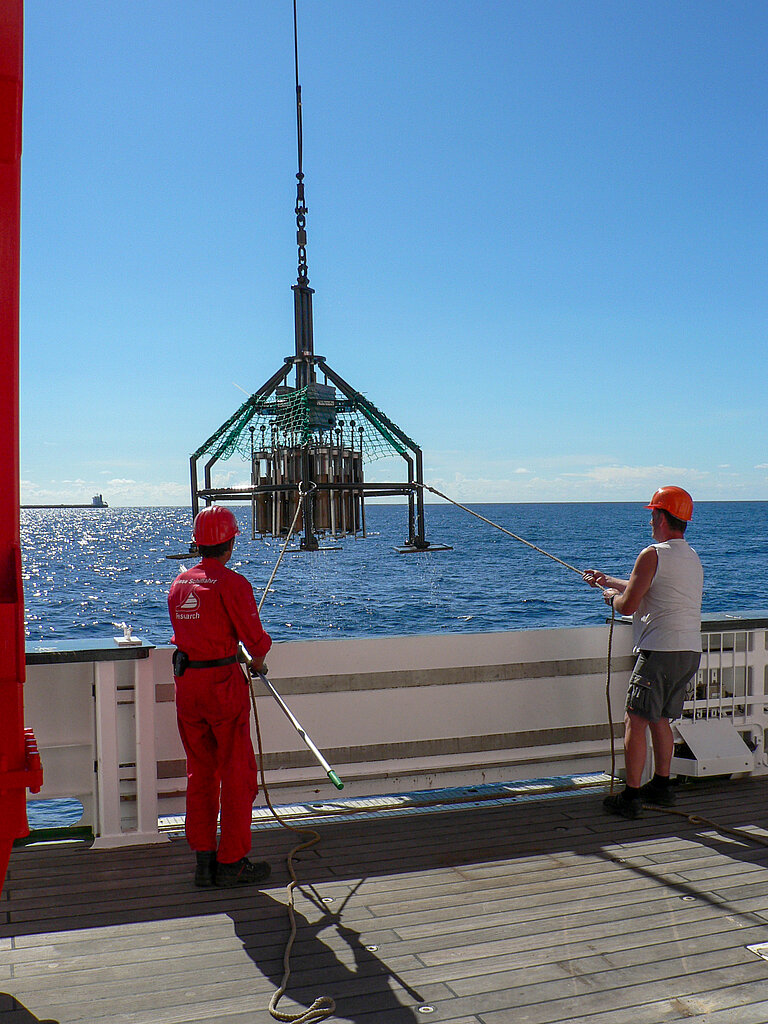  Two workers wearing red hard hats stand on board a research vessel while a piece of research equipment is hoisted on board