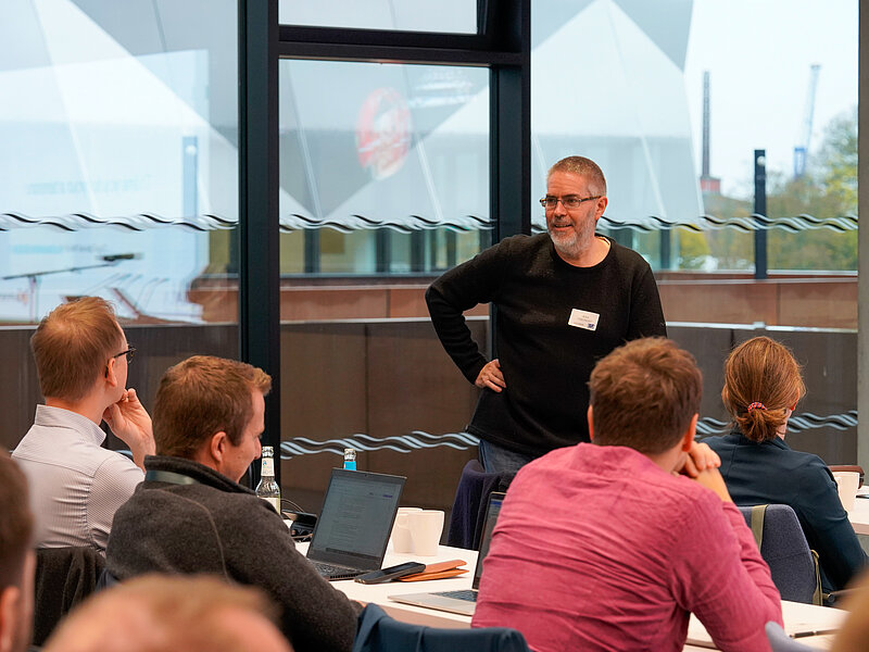 Ein Mann mit Brille und kurzen grauen Haaren spricht zu einer Gruppe von Zuhörern an Tischen in einem Konferenzraum