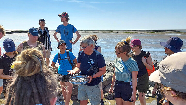 A group of young people gather in the mudflats around a woman with short white hair who is holding a bowl and explaining something.