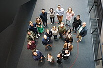 Mehrere Personen stehen auf einem dunkeln Teppich in einer Spirale angeordnet. Sie schauen nach oben zur Kamera. 