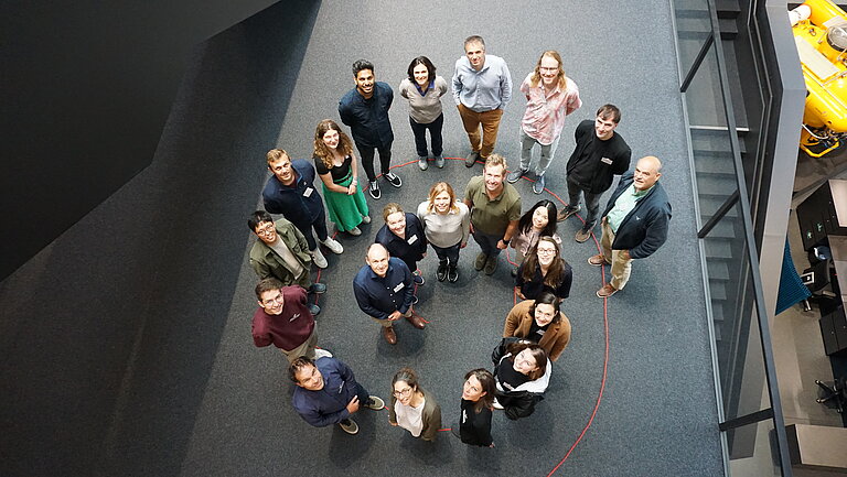 Several people are standing on a dark carpet, arranged in a spiral. They look up towards the camera. 