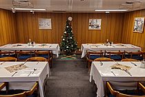 A decorated Christmas tree and four nicely set tables in the low mess hall aboard a ship.