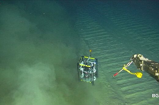 Aufnahme vom Meeresgrund, ein kleines Gestell mit Sensoren steht auf dem Grund, der Arm eines Unterwasserroboters ragt ins Bild, von links kommt eine Sedimentwolke ins Bild