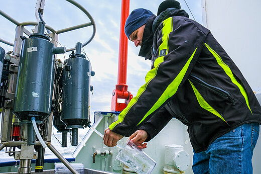 Ein Mann mit Mütze und Sicherheitsjacke lässt Wasser aus einer grauen Plastikflasche in einen Messbecher ab