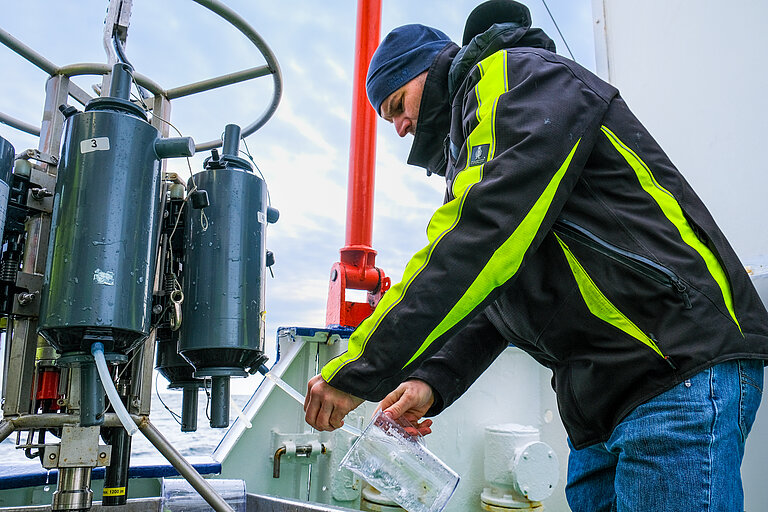 A man in a cap and safety jacket drains water from a grey plastic bottle into a measuring cup