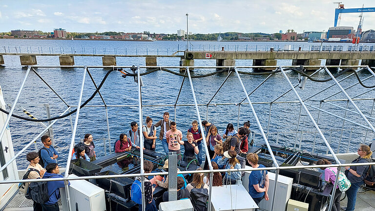  A group of young people gather around a pool of water standing on a pontoon. Water and industrial plants in the background