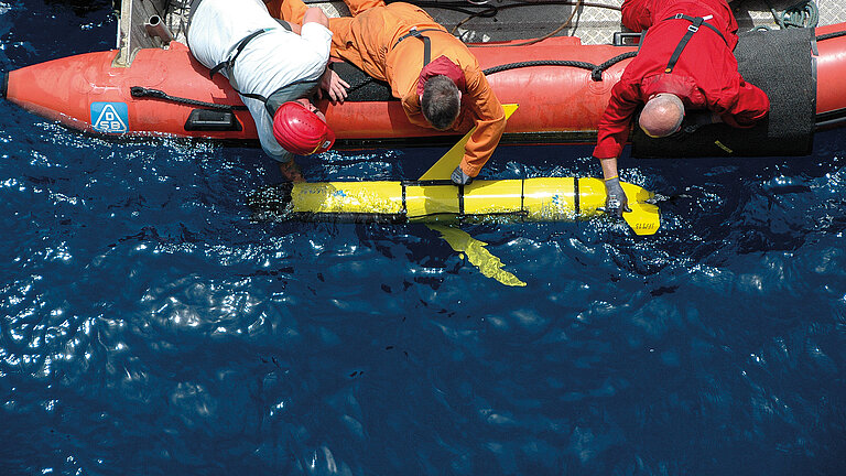  A yellow torpedo-shaped underwater measuring device is lowered into the water from a red inflatable boat