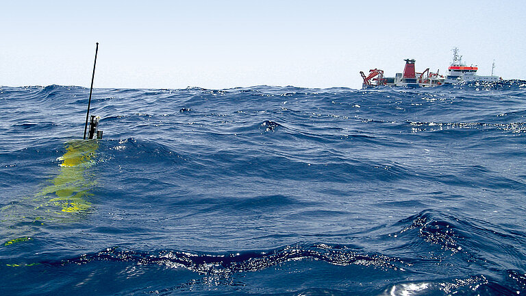 A yellow measuring buoy in the water, a research vessel on the horizon