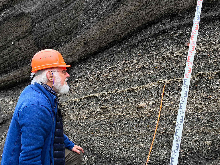  A white-haired, bearded man in an orange hard hat looks at layers of rock, next to a measuring stick