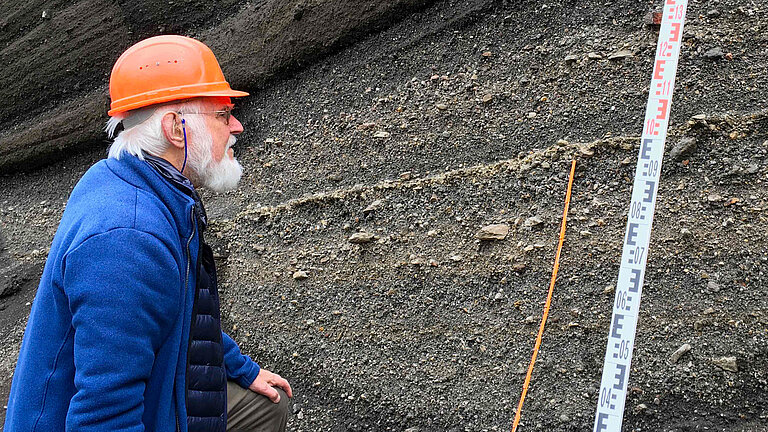 Ein weißhaariger bärtiger Mann mit einem orangefarbenen Schutzhelm schaut auf Gesteinsschichten , daneben eine Messlatte