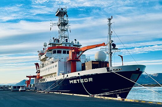 The research vessel METEOR is moored in the harbour. The sky is blue with clouds. 