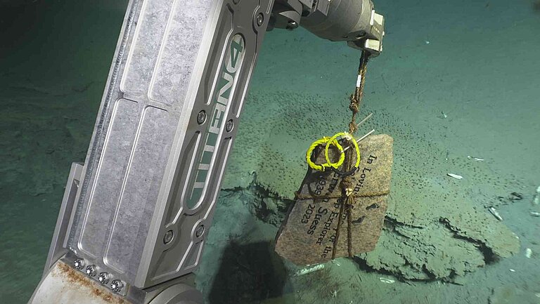 A memorial rock hangs from a robot-like arm. It is the gripper/arm of the submersible ALVIN
