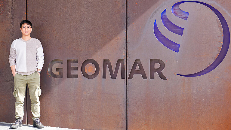 A young man with Asian features stands in front of a rust-coloured wall with the word GEOMAR written on it.