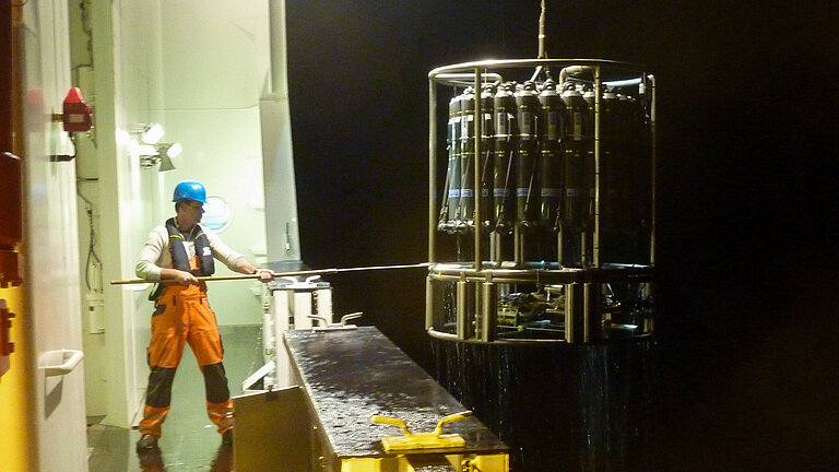 A worker in a blue hard hat uses a hook to haul a piece of research equipment back on board