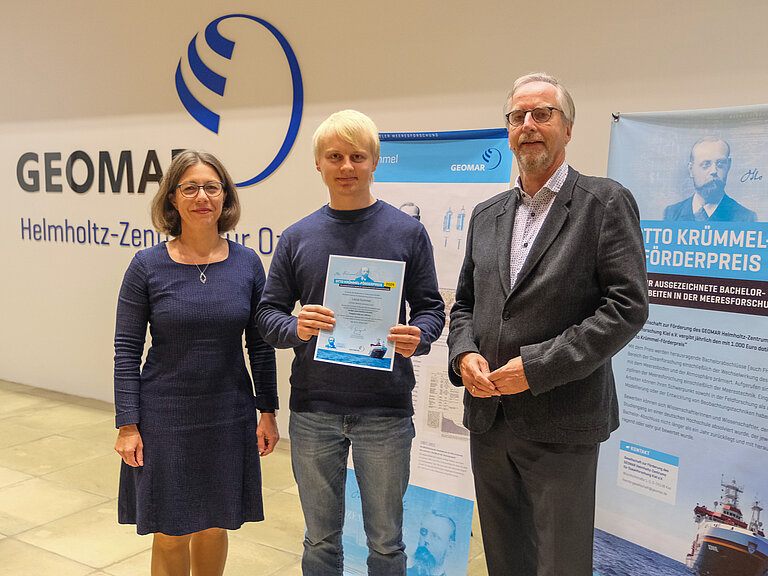 A woman and two men smile into the camera, the young man in the centre holds a certificate in his hands